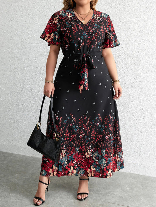 Frau in langem, schwarzem Blumenkleid mit Gürtel, hält schwarze Handtasche, trägt Sandalen.