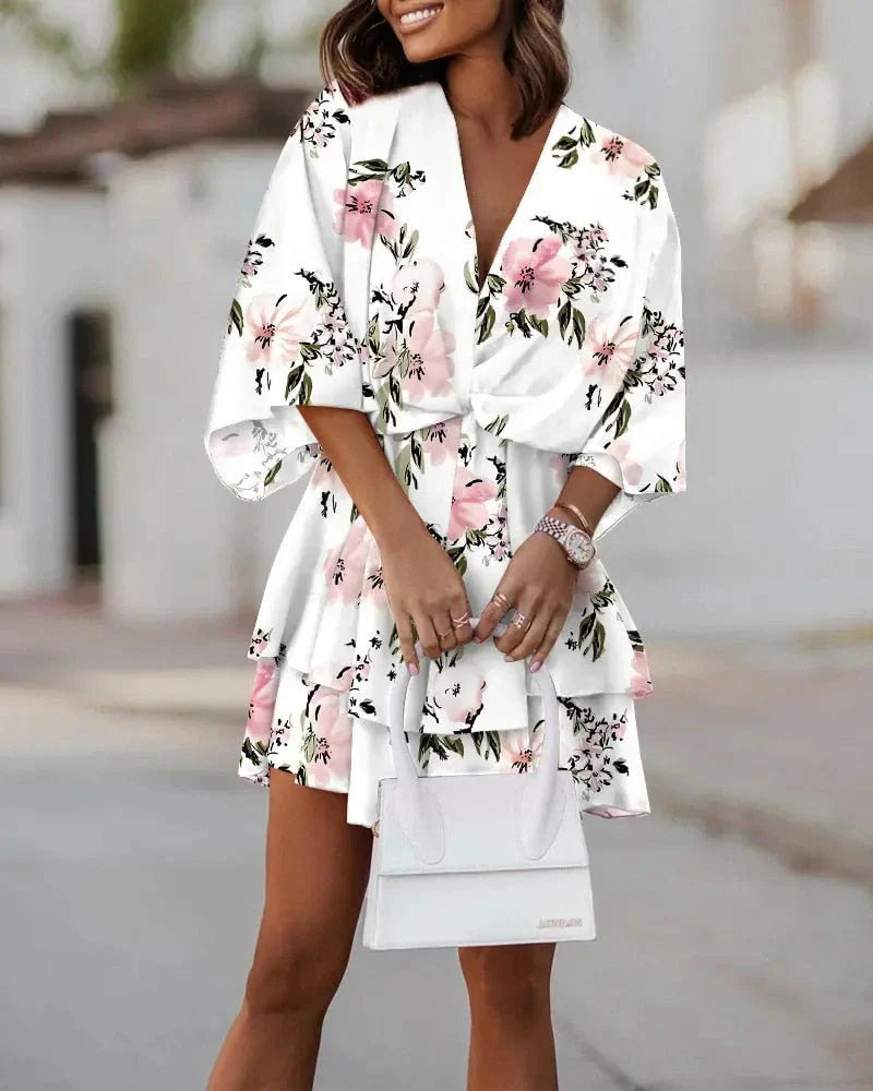 Frau in weißem, geblümtem Sommerkleid mit rosa Blumen, hält weiße Handtasche.