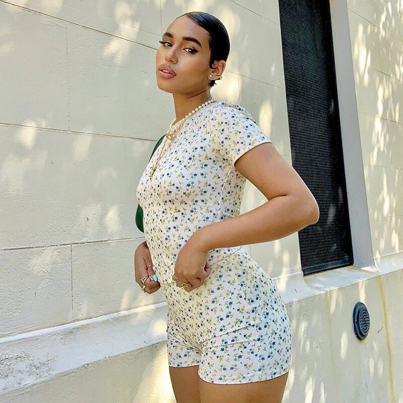 Frau in geblümtem Jumpsuit, weiß-blau, Sommermode, draußen, vor heller Wand.