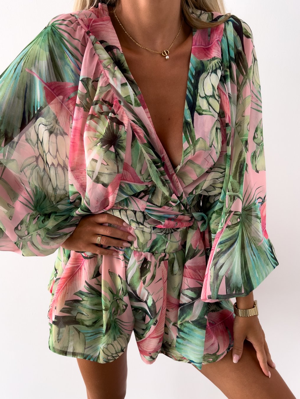 Frau in grün-rosa tropischem Jumpsuit mit Blumenmuster, V-Ausschnitt und langen Ärmeln.