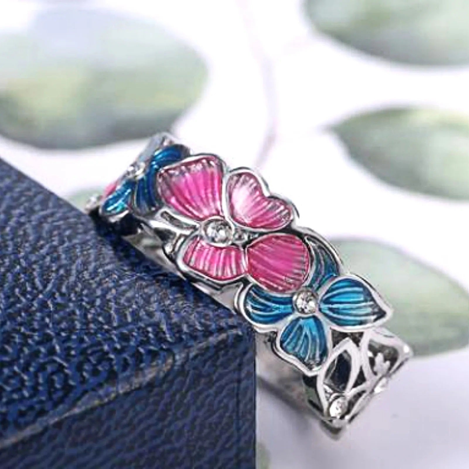 Silberring mit rosa und blauen Blumen, Kristallsteinen, auf blauem Hintergrund.