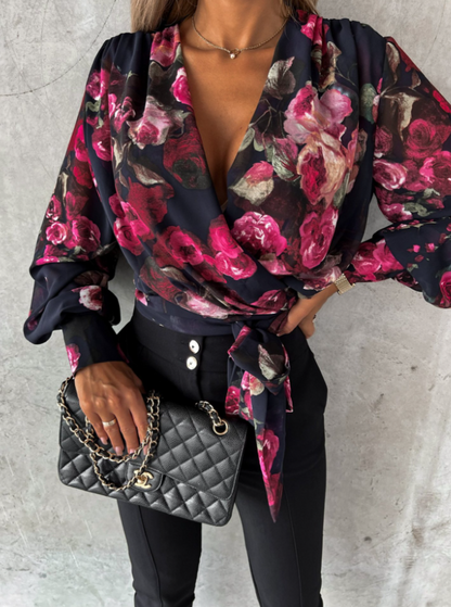 Frau in schwarzer Hose, floraler Bluse, hält schwarze gesteppte Handtasche mit Kettengriff.