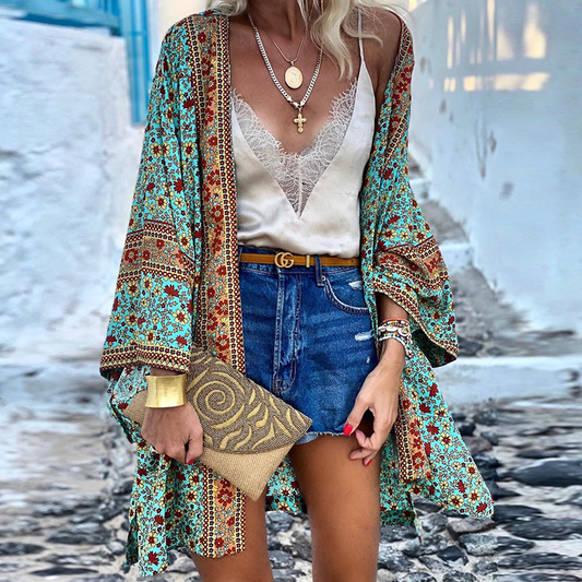 Frau in Boho-Kimono, blau-rot gemustert, mit Jeansshorts und goldener Clutch auf gepflastertem Weg.