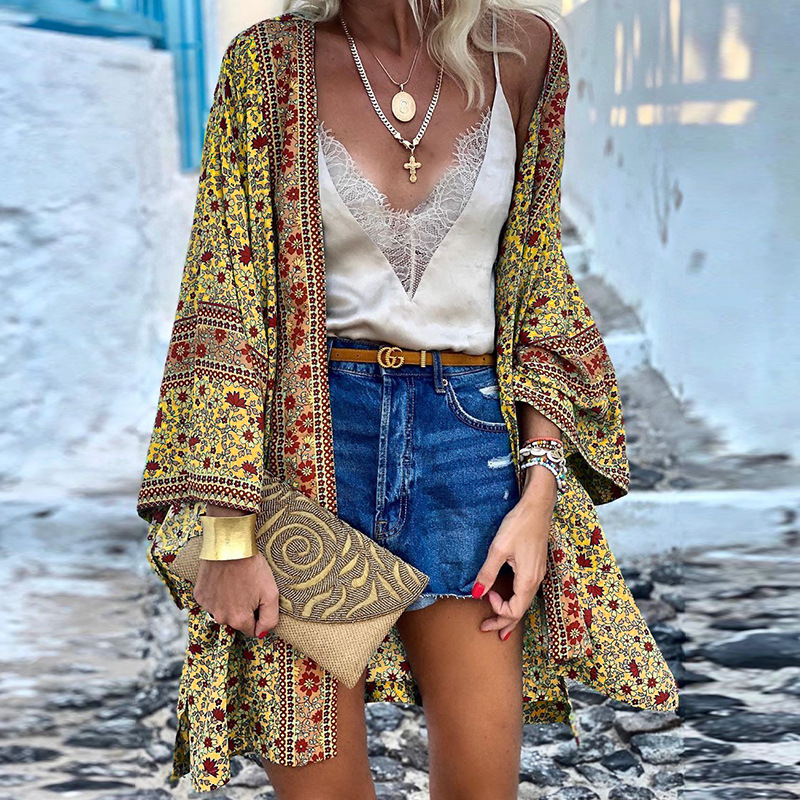 Frau in gelbem Boho-Kimono, weißem Spitzen-Top, blauer Jeansshorts, hält eine beige Clutch.