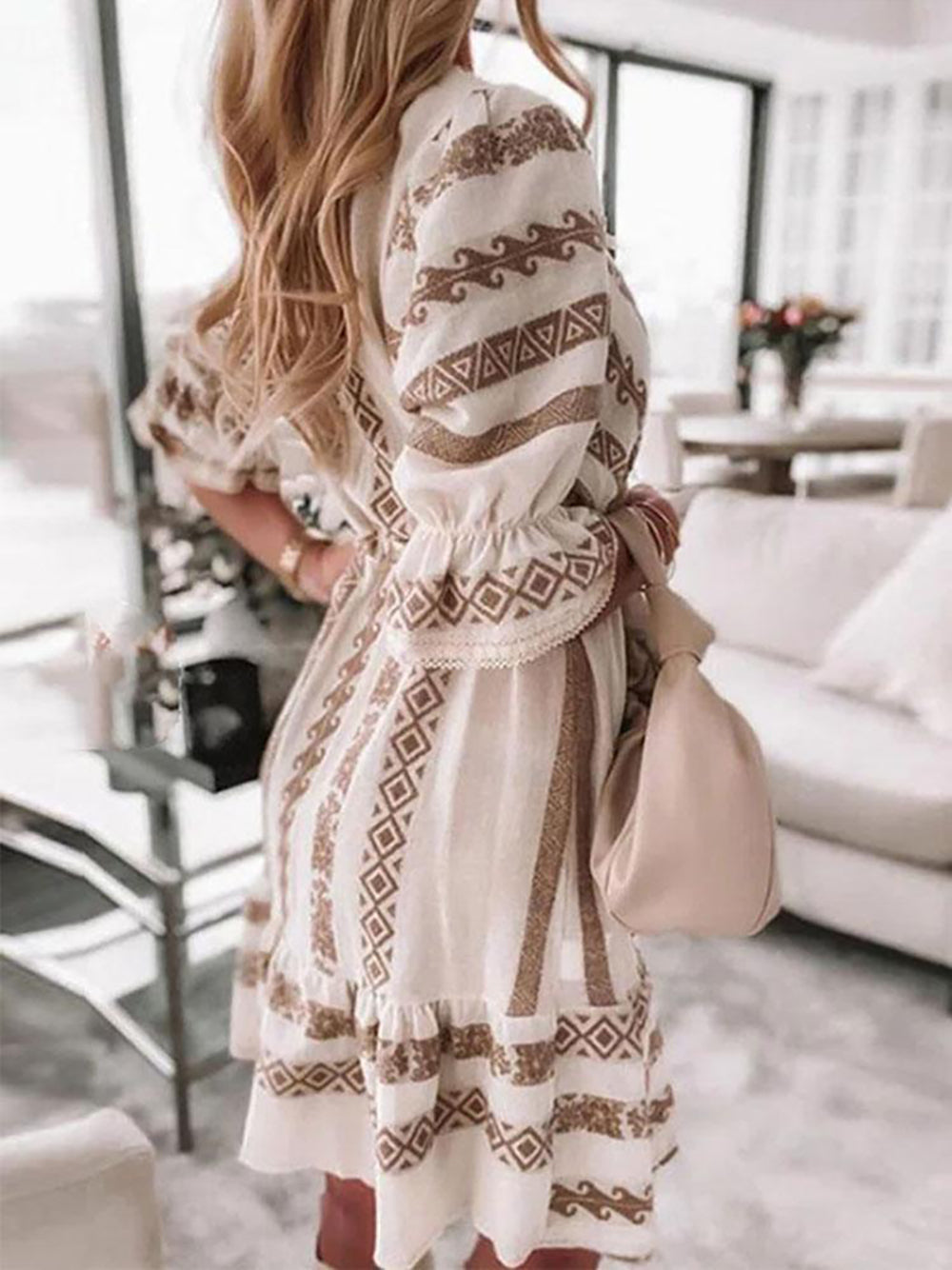 Frau in weißem Boho-Kleid mit braunen Mustern, langem Haar, in modernem Wohnzimmer.