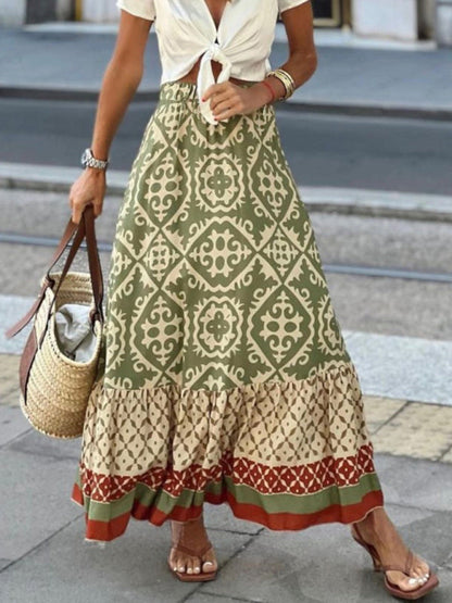 Frau in langem, grün-beigem Boho-Rock mit Ethno-Muster, trägt Strohtasche und Sandalen.