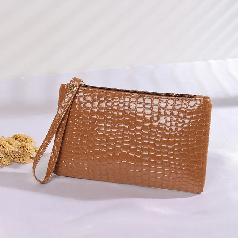 Braune Leder-Clutch mit Krokodilprägung, Reißverschluss und Handgelenkschlaufe auf weißem Hintergrund.