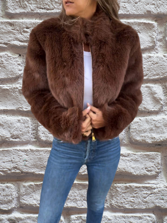 Frau in brauner Kunstpelzjacke, weiße Bluse, blaue Jeans vor Ziegelwand.
