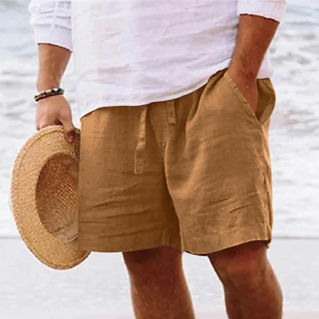 Mann in braunen Leinen-Shorts und weißem Hemd am Strand, hält Strohhut.