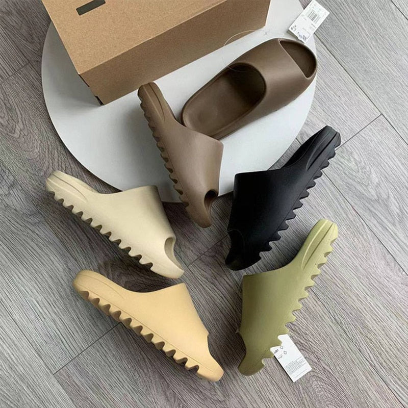 Vier Paar Yeezy Slides in Braun, Schwarz, Beige und Olivgrün auf Holzboden.
