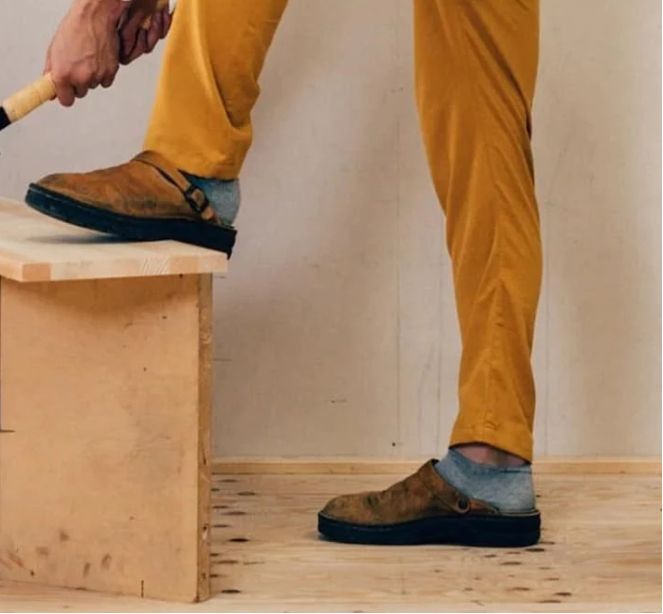 Mann in gelber Hose und braunen Wildleder-Clogs auf Holzbank, handwerkliche Arbeit.