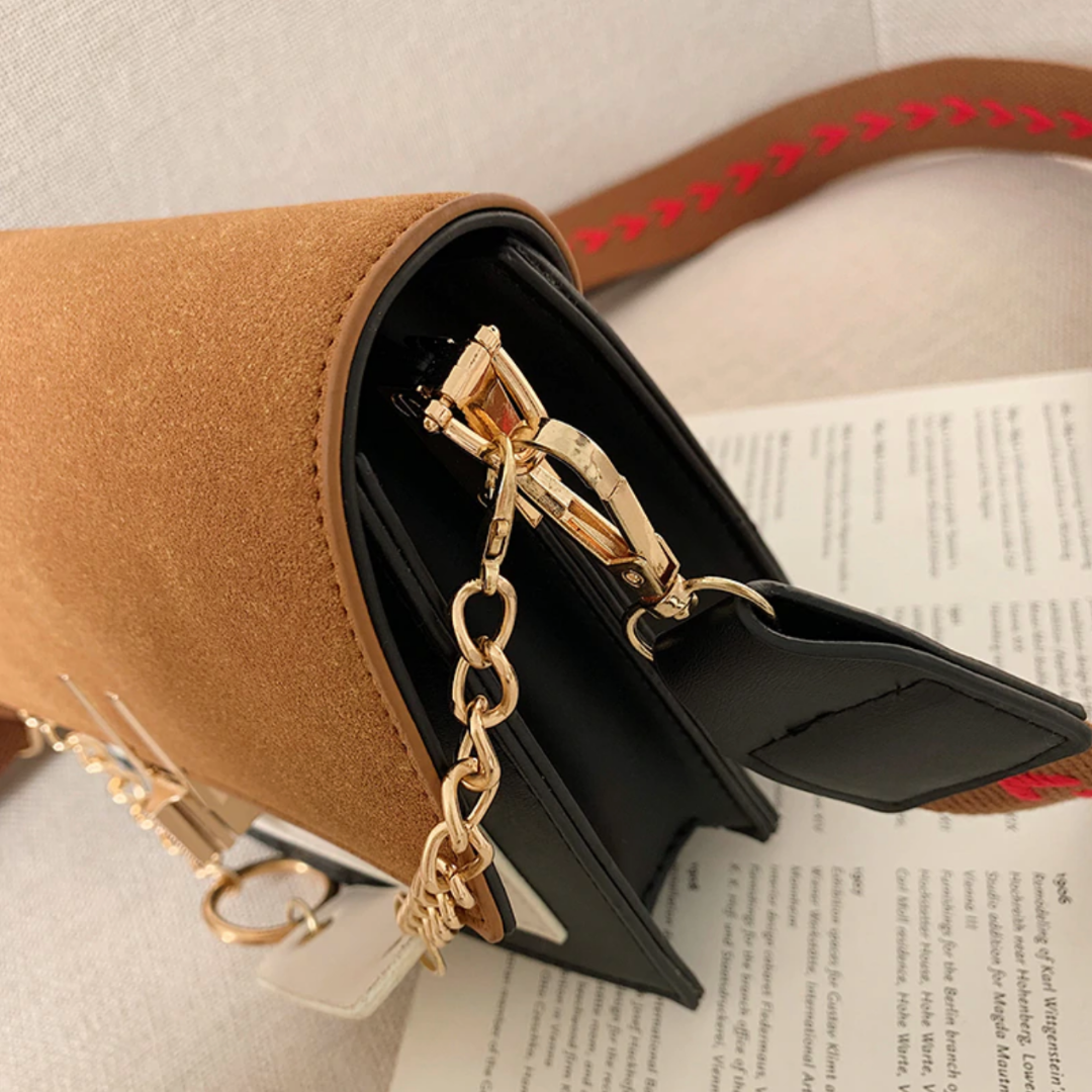 Braune Wildleder-Handtasche mit goldener Kette und schwarzem Verschluss auf Buchseite.