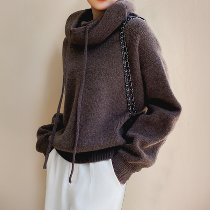 Frau trägt braunen Strickpullover mit Rollkragen und Kordeln, kombiniert mit weißer Hose.