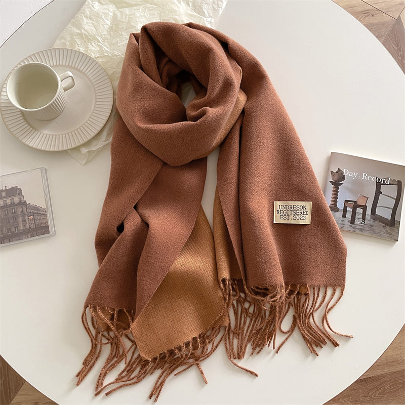 Brauner Wollschal mit Fransen auf weißem Tisch, neben Tasse und Buch, Herbstmode Accessoire.