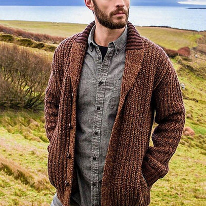 Mann in brauner Strickjacke und grauem Hemd in ländlicher Landschaft.
