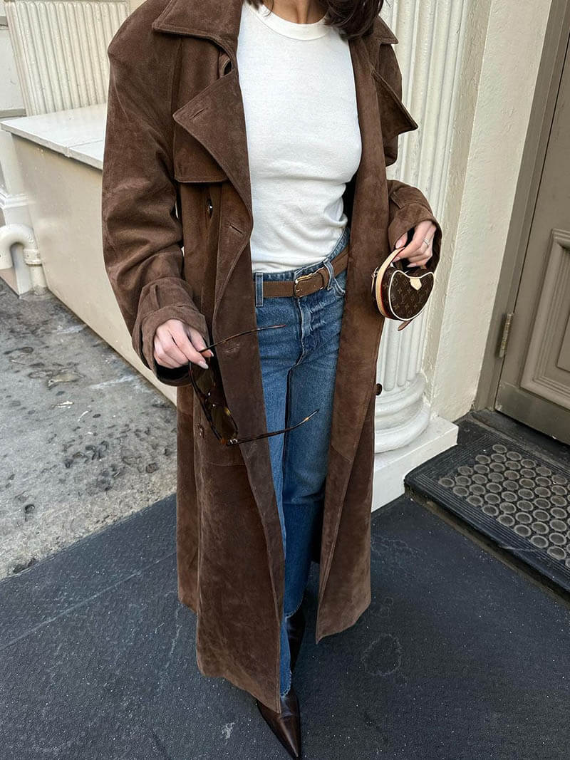 Frau in braunem Wildledermantel, weißem Shirt, blauer Jeans, hält Sonnenbrille und Herz-Tasche.