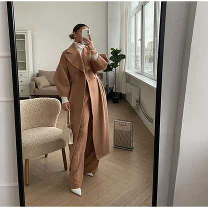 Frau in braunem Wollmantel, Anzughose, weiße Schuhe, Spiegel-Selfie, modernes Wohnzimmer.