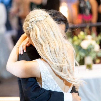 Braut mit blonder Hochsteckfrisur und Kristall-Haarband umarmt Bräutigam bei Hochzeit.