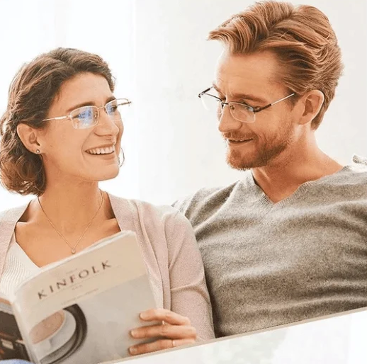 Zwei Personen mit Brillen lesen ein Magazin auf einem Sofa, lächelnd und entspannt.