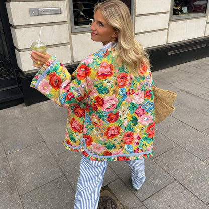Frau in bunter Blumenjacke, gestreifte Hose, mit Getränk, auf Gehweg, Sommermode.