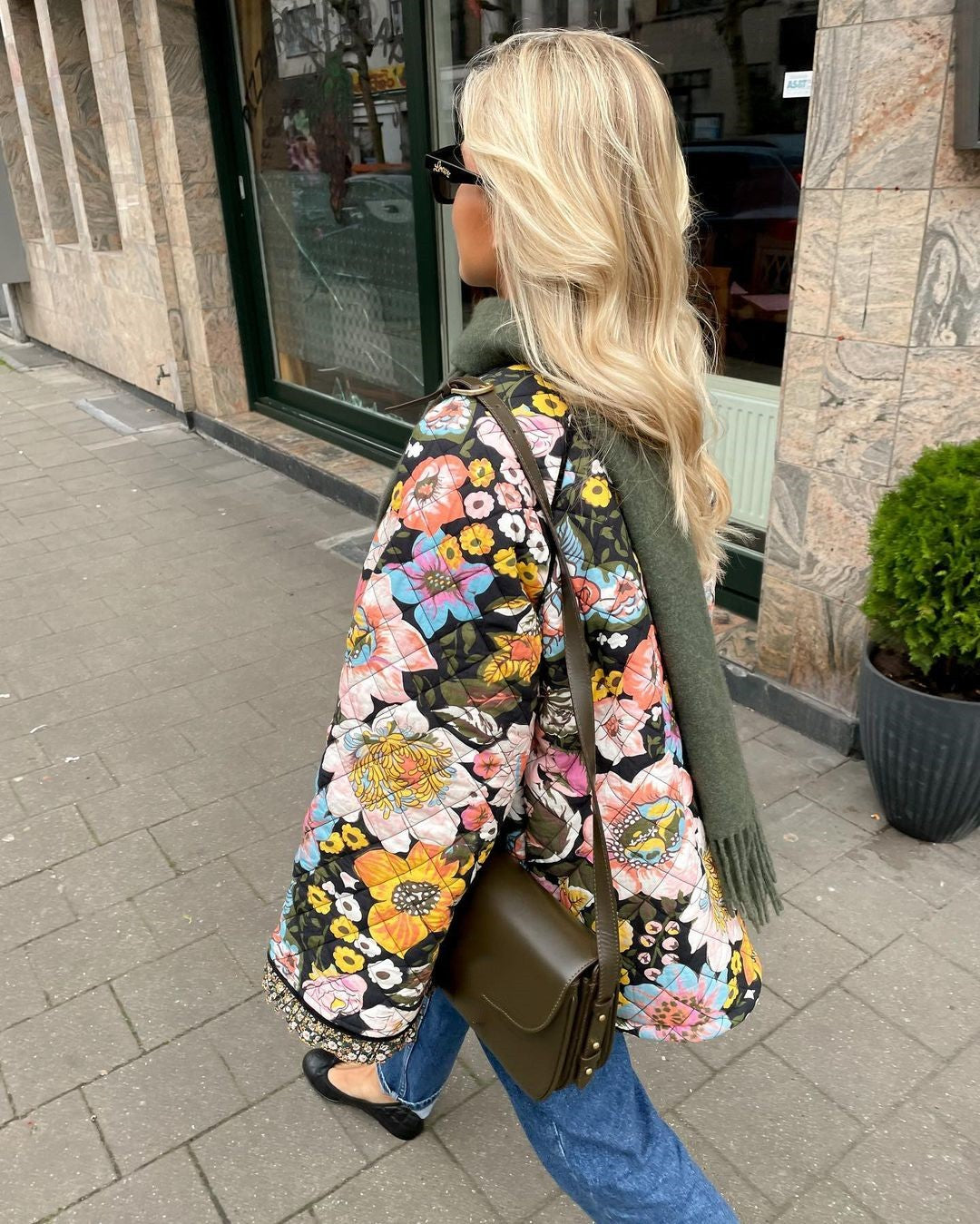 Frau mit bunter Blumenjacke, grüner Tasche und Schal spaziert auf Bürgersteig.