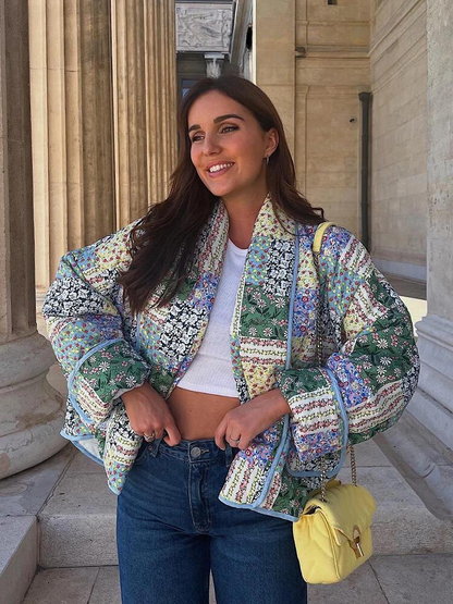 Frau in bunter Patchwork-Jacke, weißem Top, blauer Jeans, gelber Tasche, lächelnd vor Säulen.