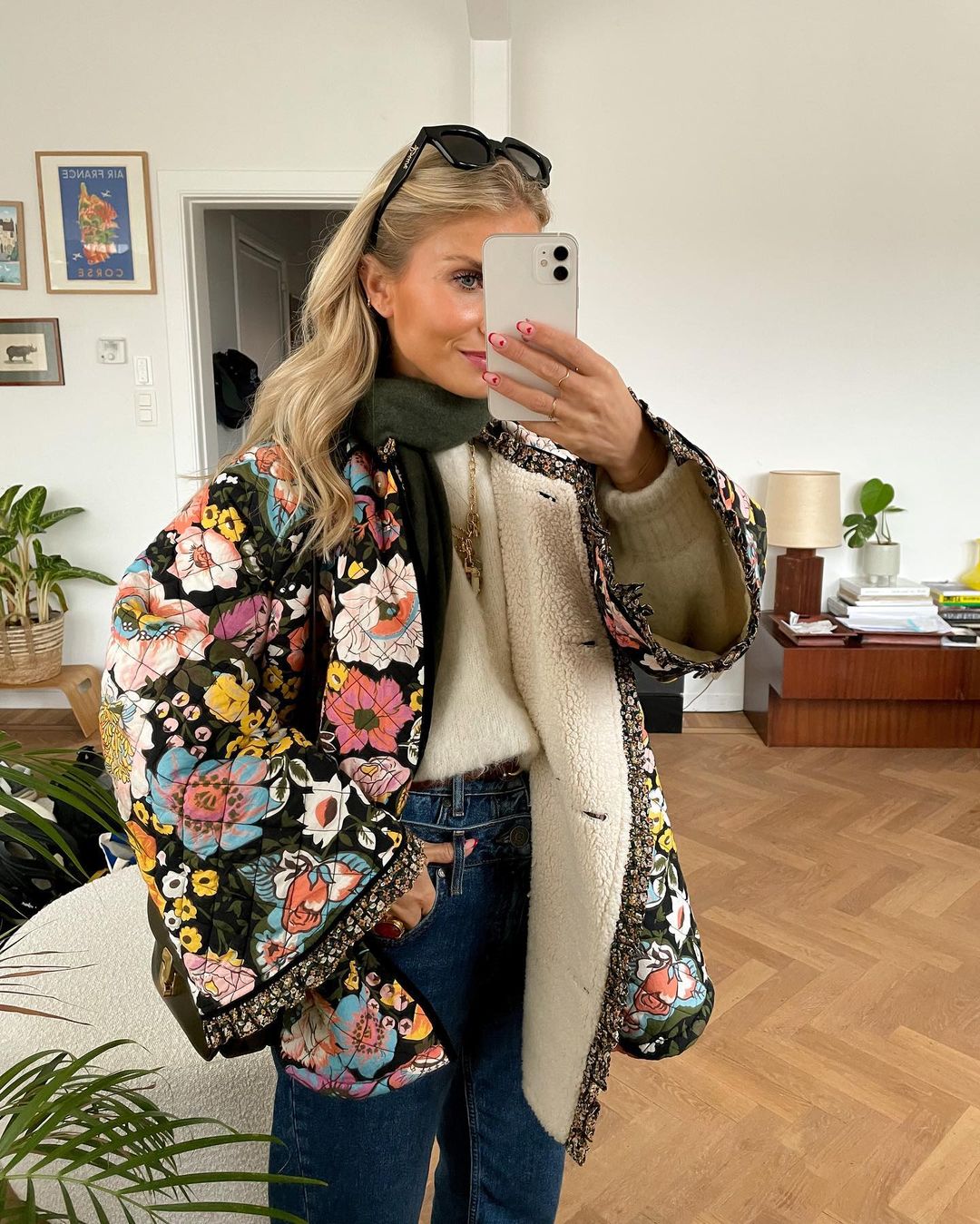 Frau in bunter Blumenjacke, weißem Pullover, blauer Jeans, macht Selfie im Wohnzimmer.