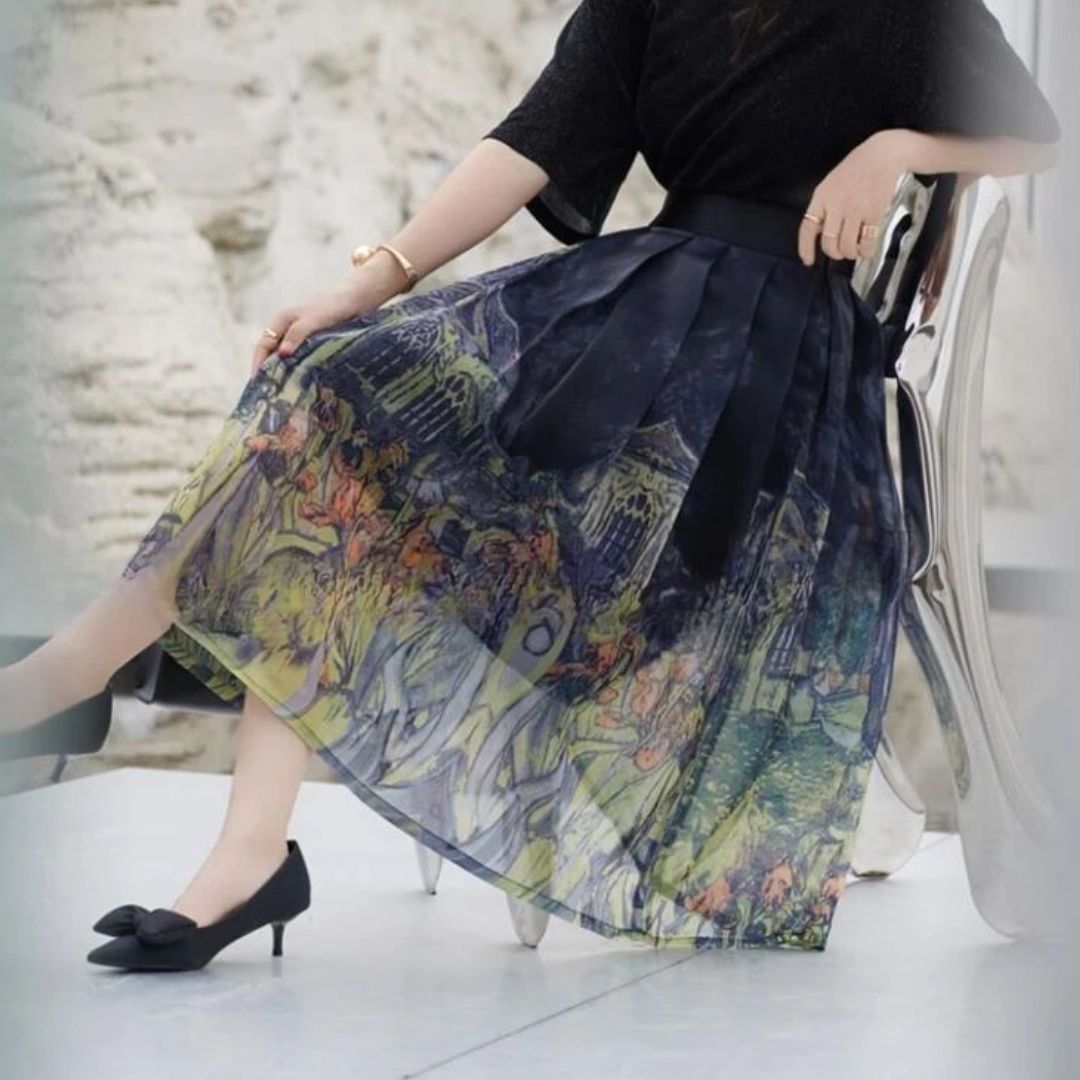 Frau in schwarzem Oberteil und bedrucktem Faltenrock, sitzt auf Stuhl, elegante schwarze Schuhe.