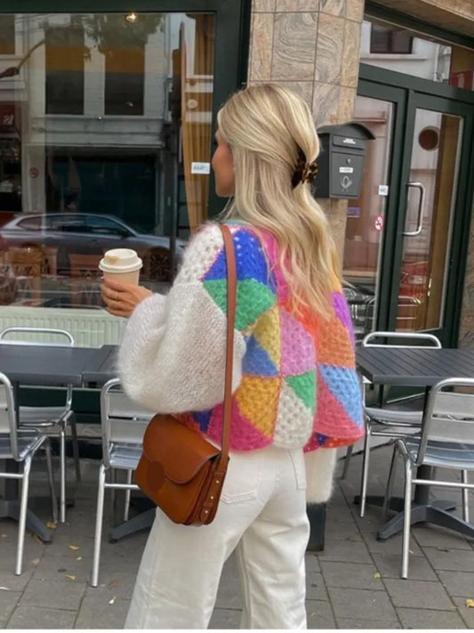 Frau mit buntem Strickpullover, weißer Hose, brauner Tasche, Kaffeebecher, draußen, Stadt.