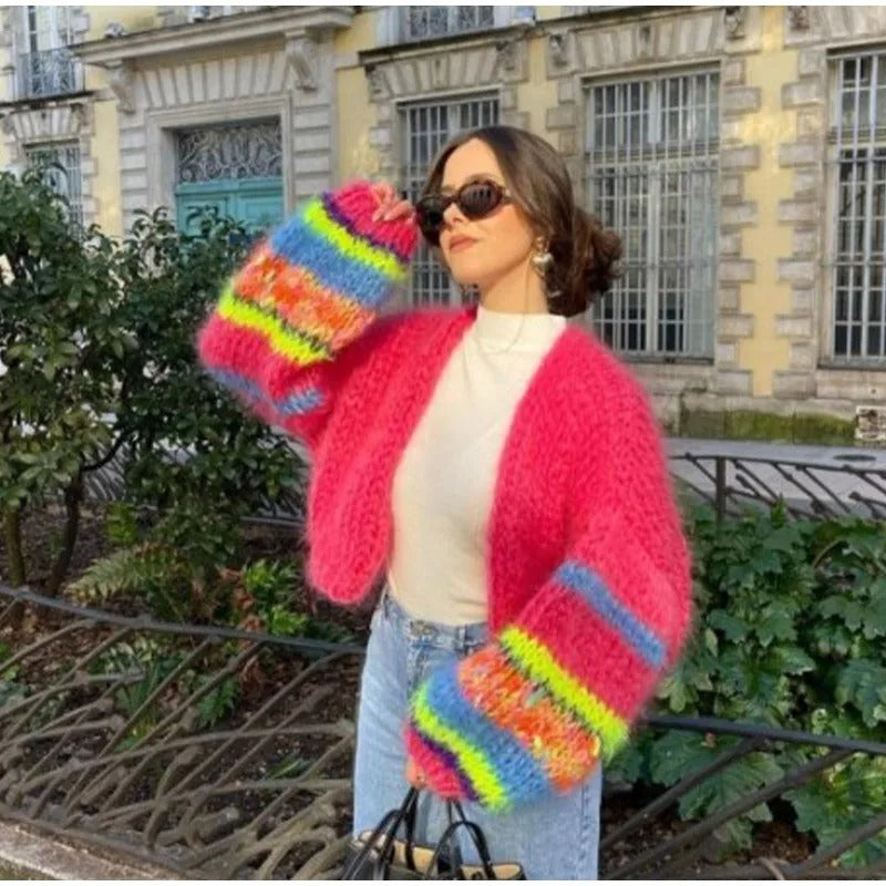 Frau in pinker Strickjacke mit bunten Ärmeln, Sonnenbrille, draußen vor Gebäude.