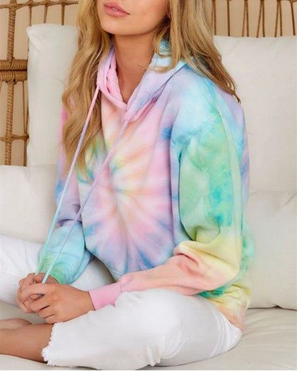 Frau in pastellfarbenem Batik-Hoodie, sitzt auf weißem Sofa, lässige Mode, gemütlicher Stil.