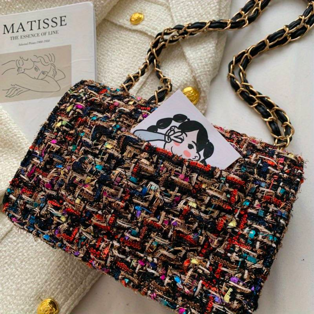 Bunte Tweed-Handtasche mit Kettengurt auf weißem Stoff, Matisse-Buch im Hintergrund.