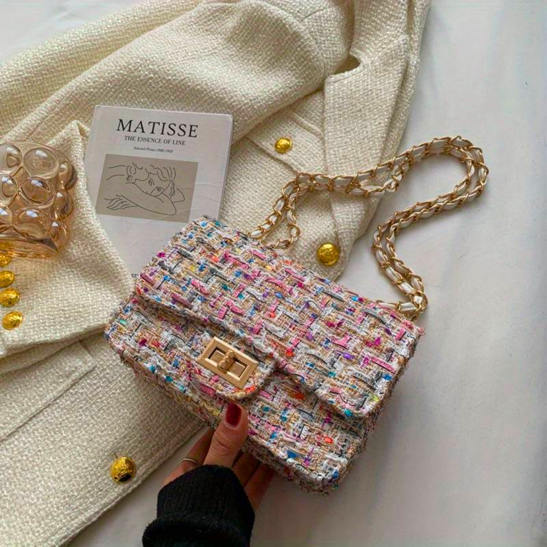 Bunte Tweed-Handtasche mit Kettengurt auf weißem Stoff, neben Matisse-Buch und Glas.