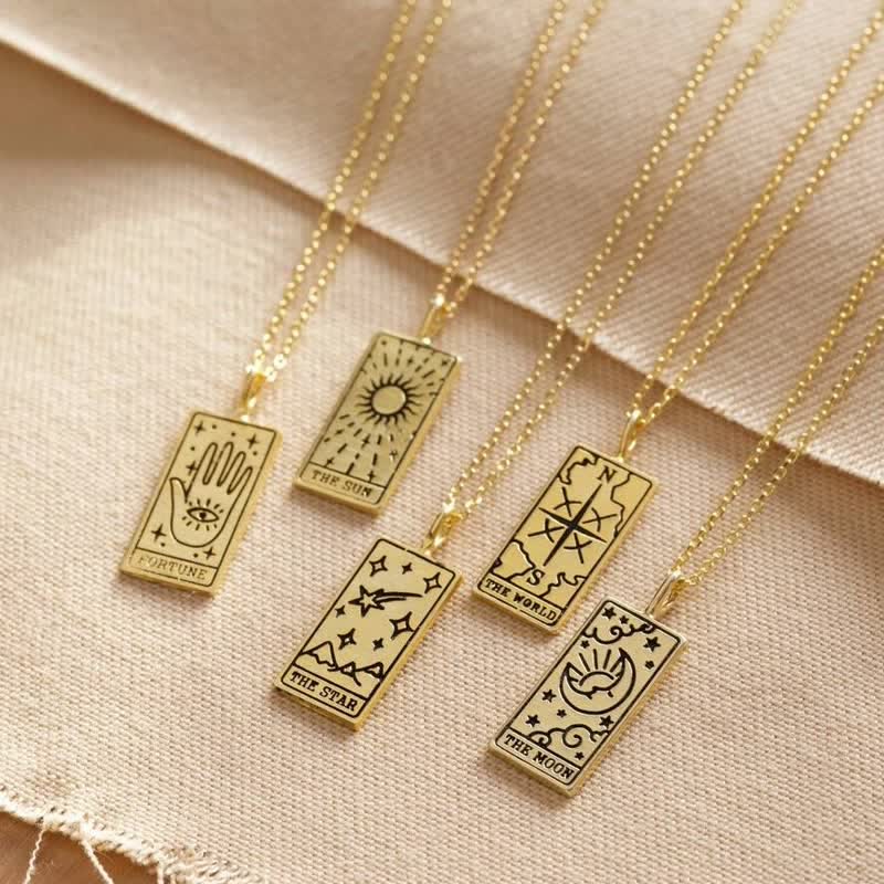 Fünf goldene Tarot-Anhänger auf Ketten, Fortune, Sun, World, Star, Moon, auf beigem Stoff.
