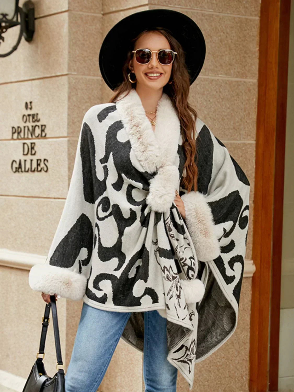 Frau in grauem Poncho mit schwarzem Muster, Pelzkragen, Hut und Sonnenbrille vor Hotel.