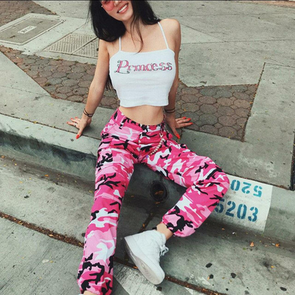 Frau in rosa Camouflage-Hose und weißem "Princess"-Top, sitzt auf Bürgersteig.