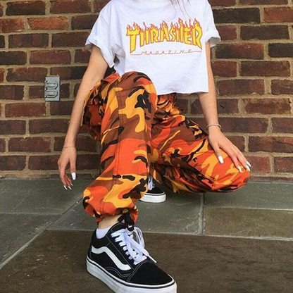 Frau in Thrasher-Shirt und orange Camouflage-Hose, kniend vor Ziegelwand, trägt Vans-Sneaker.