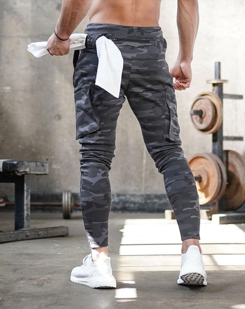 Mann in grauer Camouflage-Trainingshose im Fitnessstudio, hält weißes Handtuch, Hanteln im Hintergrund.