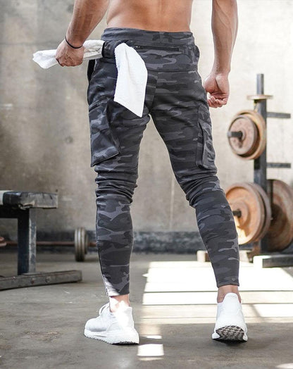Mann in grauer Camouflage-Trainingshose im Fitnessstudio, hält weißes Handtuch, Hanteln im Hintergrund.