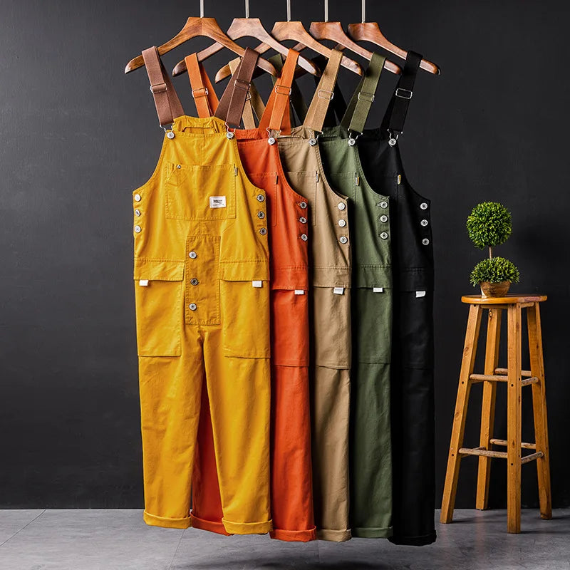 Fünf Herren-Latzhosen in Gelb, Orange, Beige, Grün, Schwarz auf Kleiderbügeln vor dunklem Hintergrund.