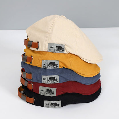 Stapel von Schiebermützen in Beige, Gelb, Blau, Rot, Schwarz mit Lederriemen und Etikett.