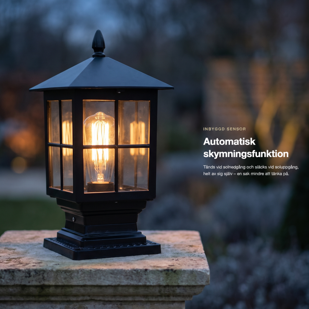 Tänd svart pelarlampa på en stenpelare i skymningen, med suddig trädgård i bakgrunden och en ruta med text om automatisk tändning. 2