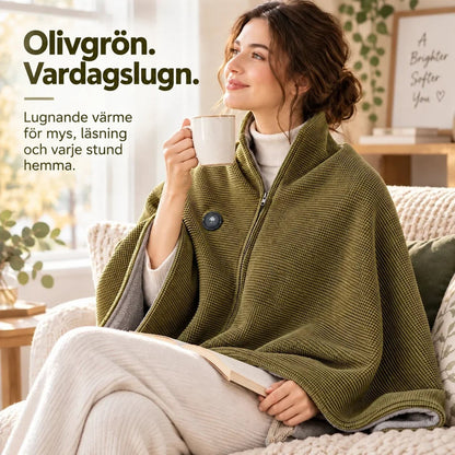 Kvinna i olivgrön poncho över axlarna sitter i soffan med en bok och en kopp 8