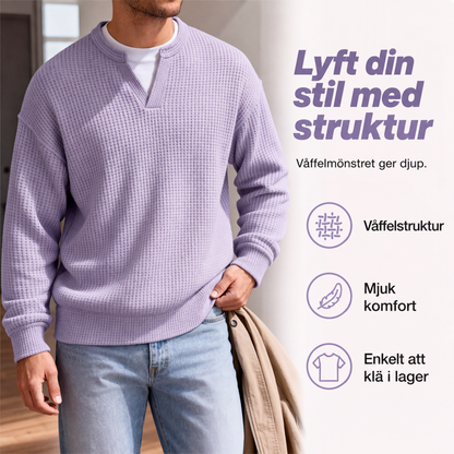 Närbild på lila våffelstickad topp med V-ringning över vitt linne, buren av man med beige jacka över armen. 3