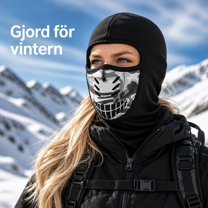Kvinna med svart balaklava och kamouflagemönstrad ventilerad munskydd poserar i en snöig bergsmiljö, klädd i svart jacka och ryggsäck. 1