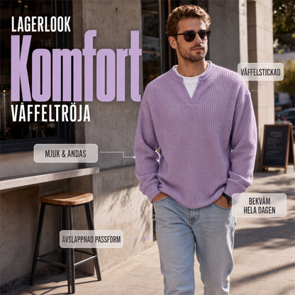 Man i sjalviolett våffelstickad topp och ljusa jeans går utomhus på en gata, med solglasögon och handen i fickan. 1