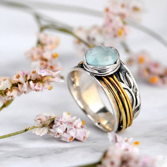 Silberring mit Aquamarin-Stein und goldenen Akzenten, umgeben von rosa Blumen auf Marmor.