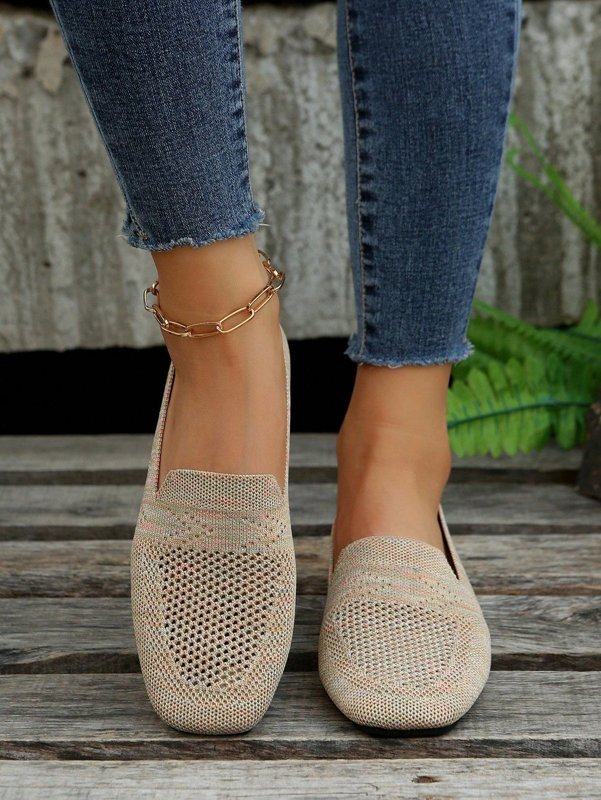 Beige slip-on-skor i luftigt mesh med fyrkantig tå sedda framifrån på en modell i blå jeans, med trädäck och ormbunke bakom. 3