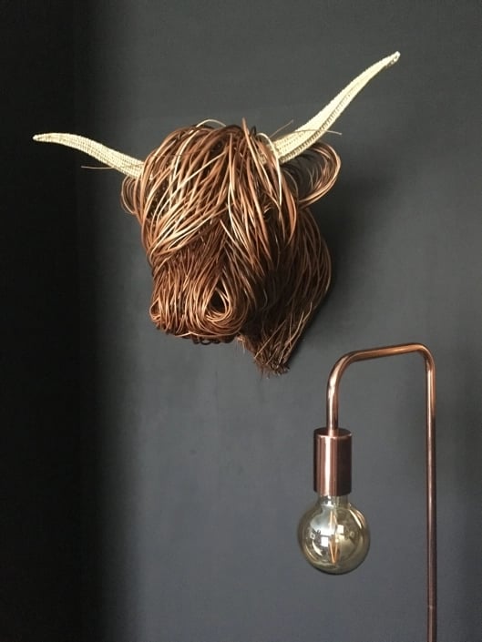 Flätad kohuvudskulptur med ljusa horn på mörkgrå vägg bredvid en kopparfärgad lampa 1