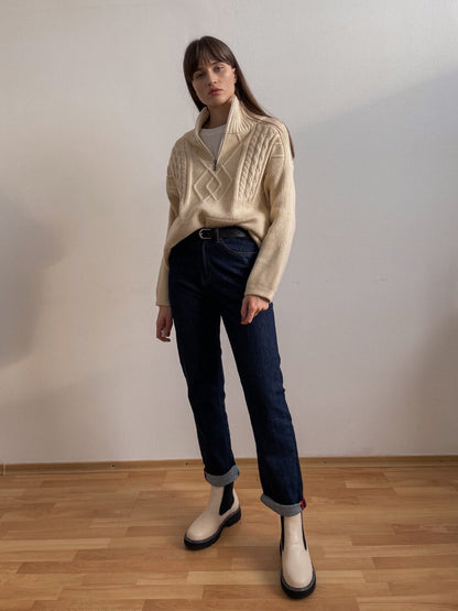 Frau in cremefarbenem Strickpullover und blauen Jeans, weiße Stiefel, modischer Look.